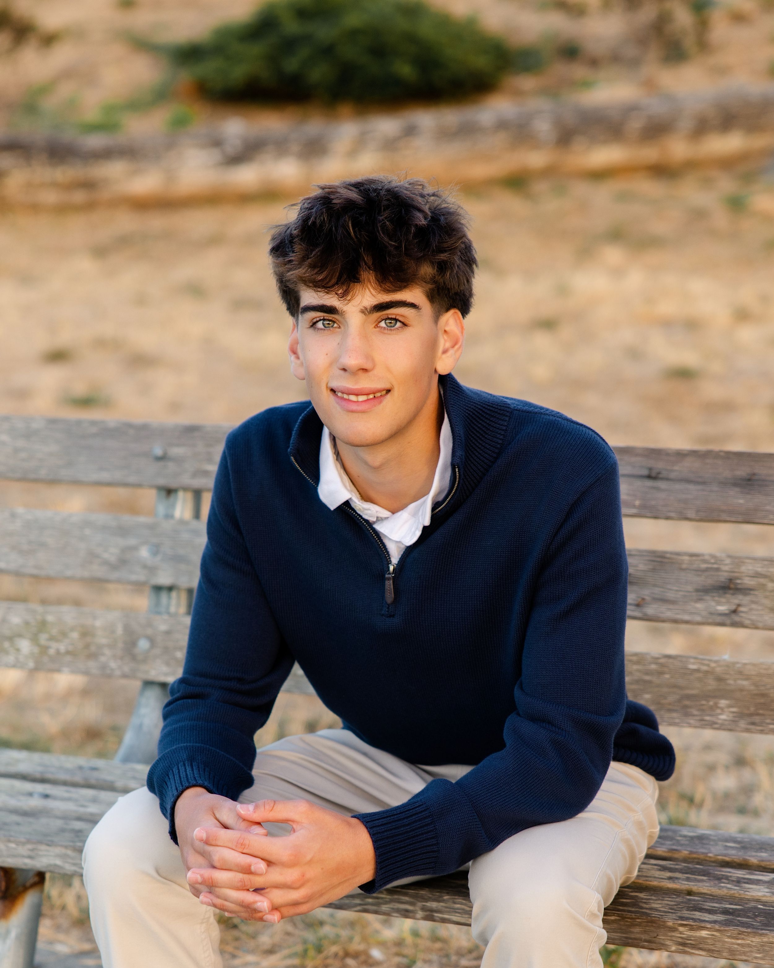 Young man in navy sweater sitting on wooden bench outdoors, smiling confidently in a casual senior portrait photo.