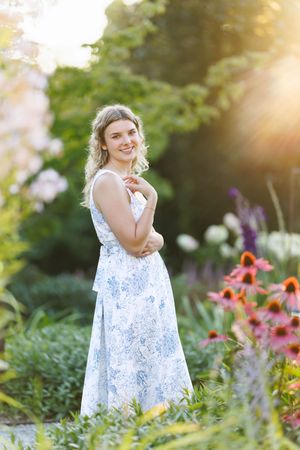 Professional senior portrait. Smiling blonde woman in floral maxi dress posing in a colorful sunlit flower garden.