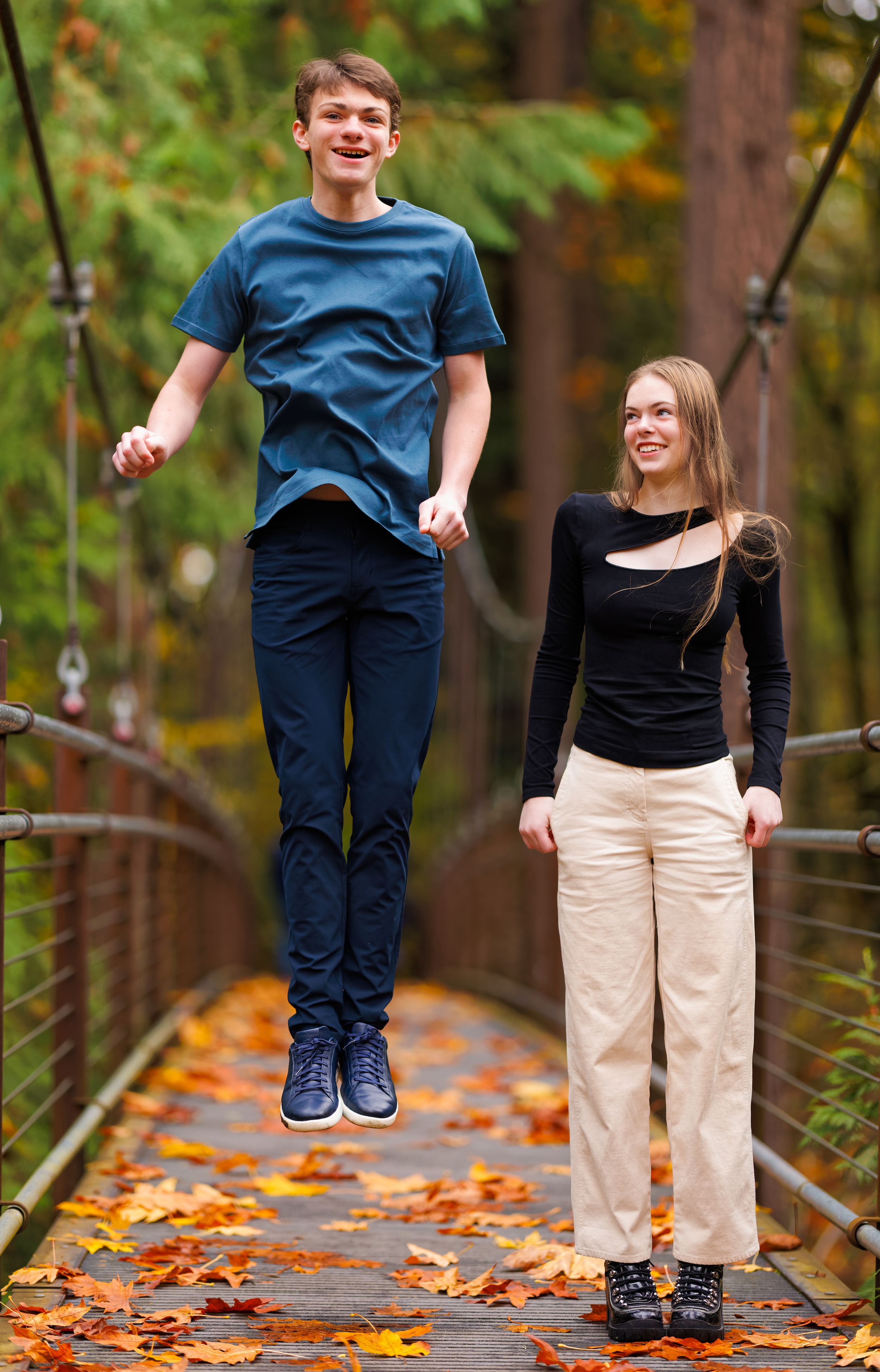 Teen boy jumping on a wooden suspension bridge beside a smiling girl, surrounded by colorful autumn leaves.