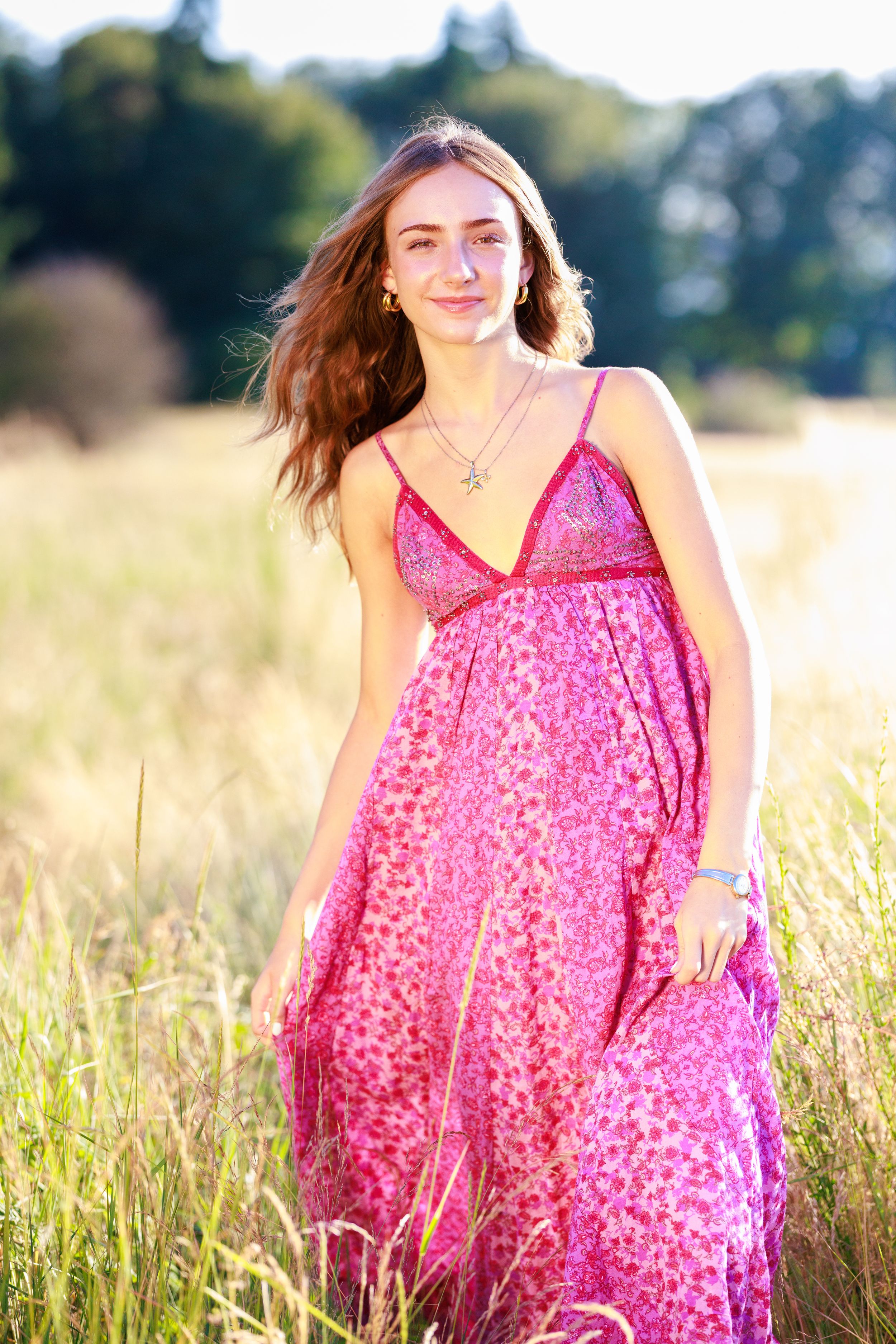 Young woman in pink floral maxi dress standing in sunlit field with long brown hair flowing in breeze.