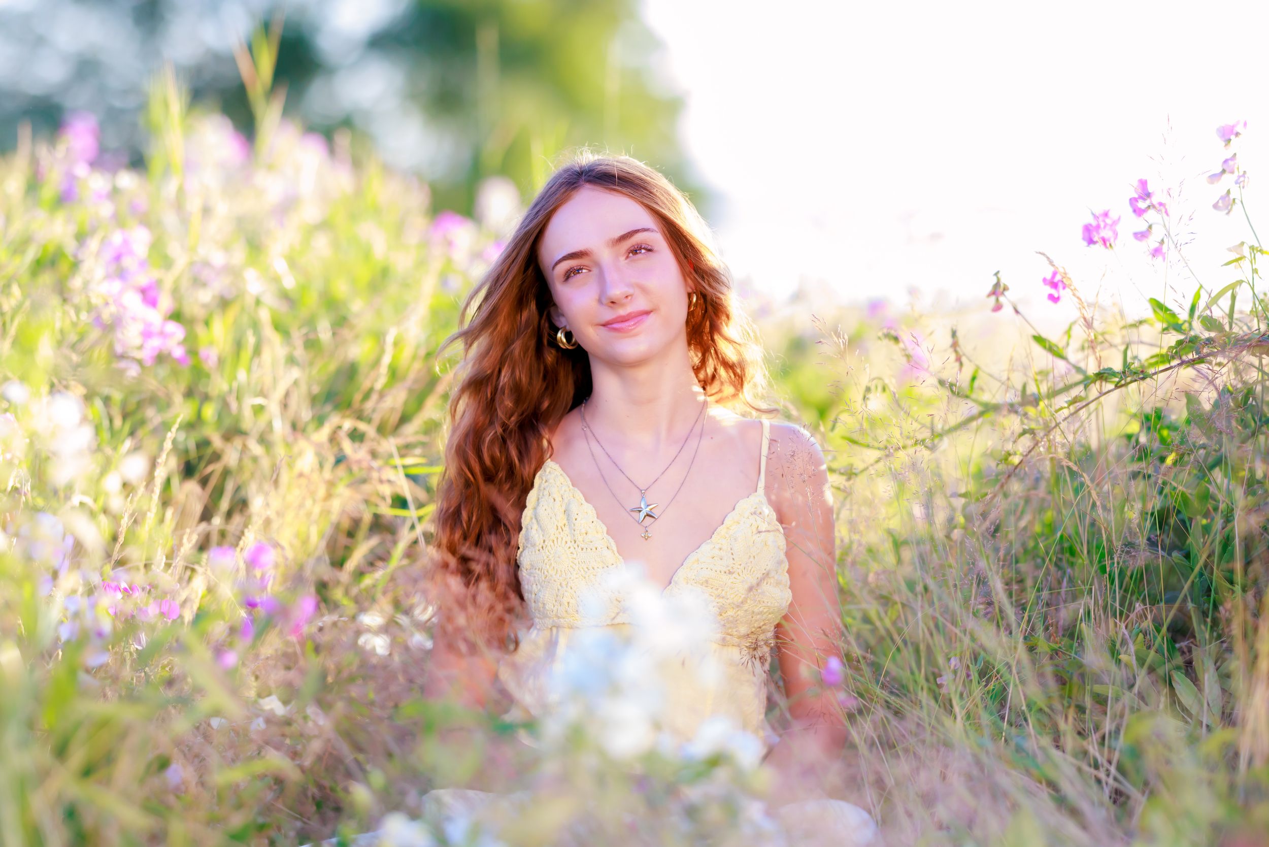 Young woman with red hair in yellow dress sitting among wildflowers in a sunlit meadow.
