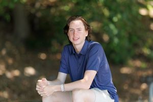 Young man in blue polo shirt sitting outdoors among trees, smiling casually in a relaxed portrait setting.