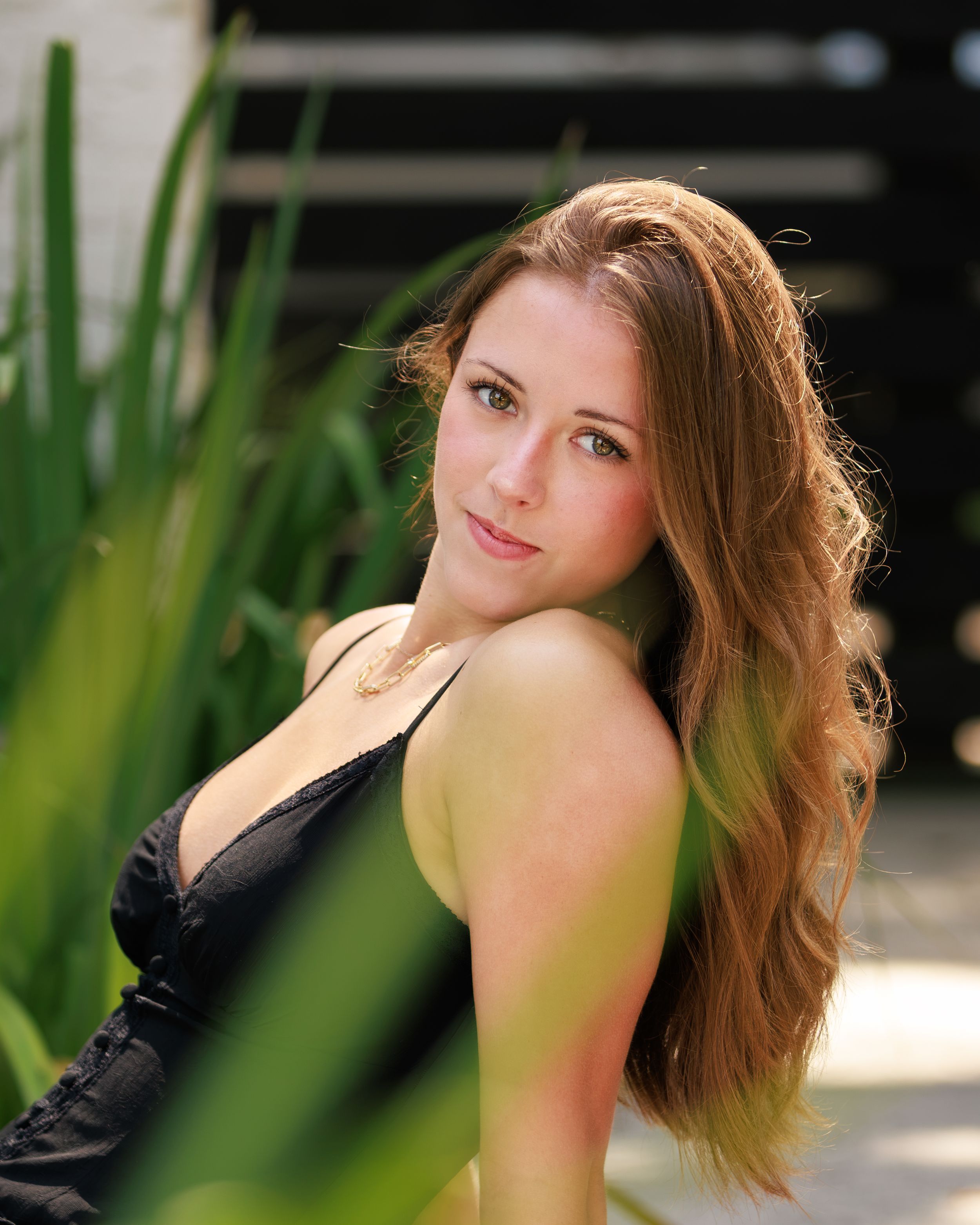 Young woman with long auburn hair poses outdoors among green plants, wearing a black top in natural sunlight. Professional senior portrait.