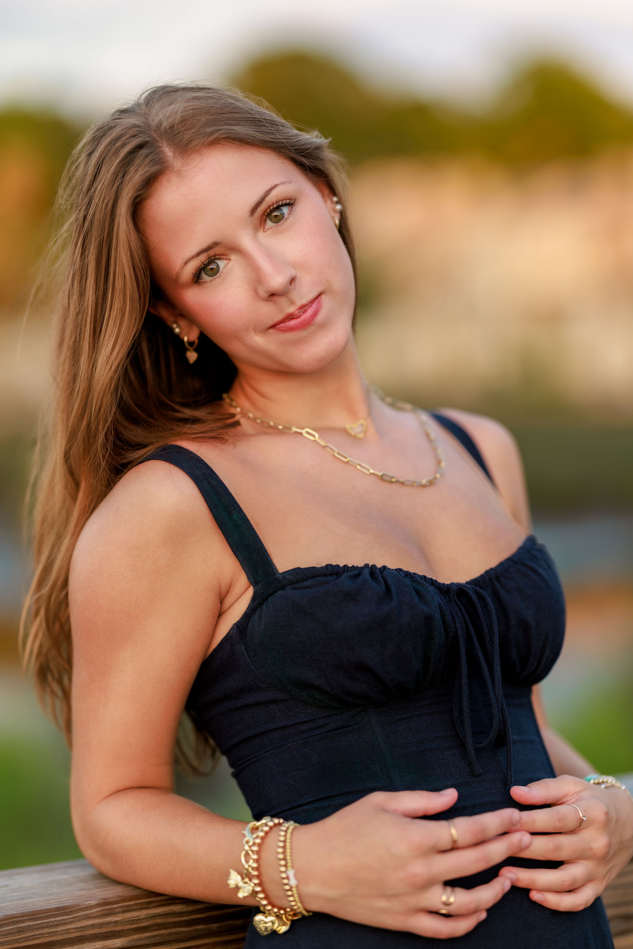 Young woman in black dress with gold jewelry poses outdoors at golden hour, long blonde hair, green eyes.