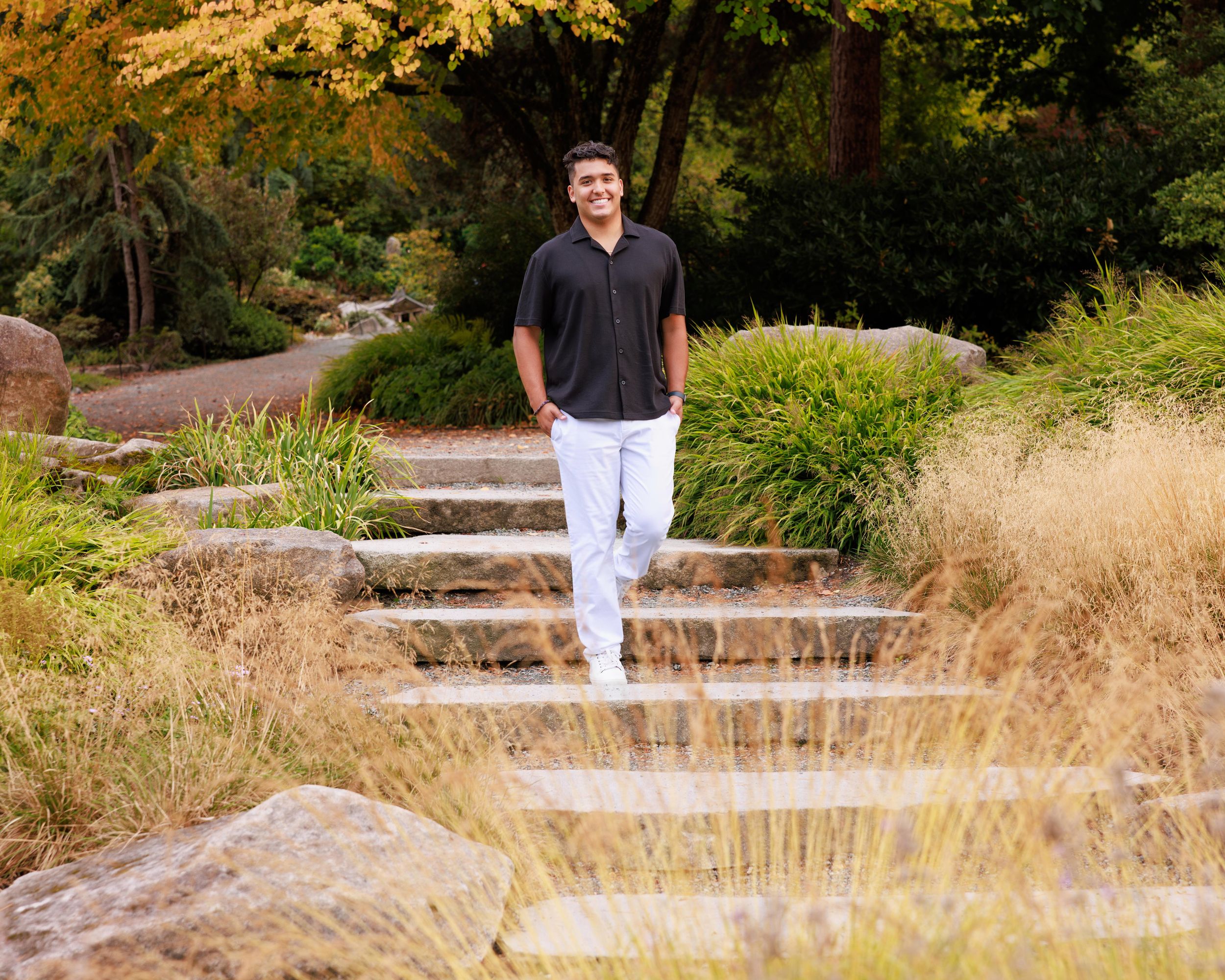 Young man in black polo and white pants standing on stone steps surrounded by autumn foliage in a garden park.