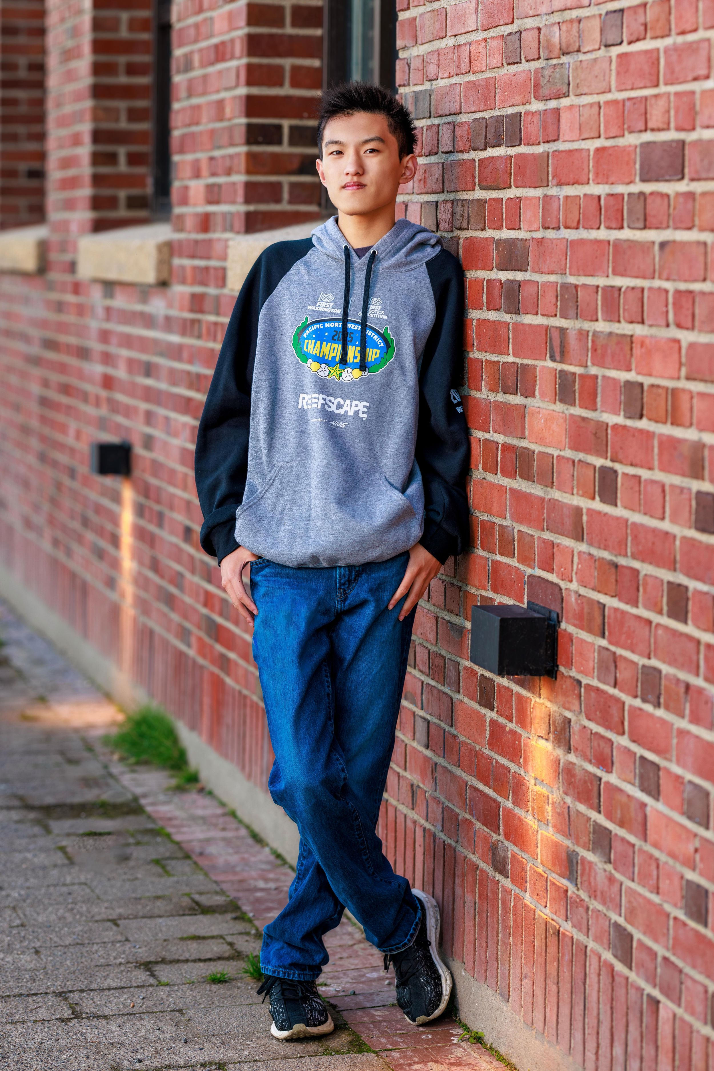 Teen boy in gray hoodie and jeans leaning against a red brick wall outdoors.