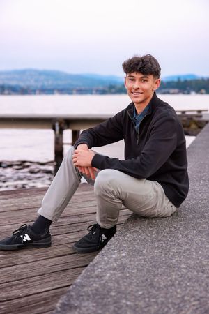 Smiling teenage boy sitting on a wooden pier by the water, wearing a black jacket and gray pants with black sneakers.