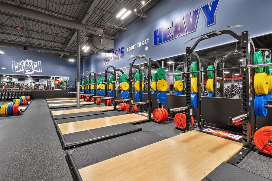 Modern gym with rows of barbells, weight racks, and colorful plates on wooden platforms under blue signage.