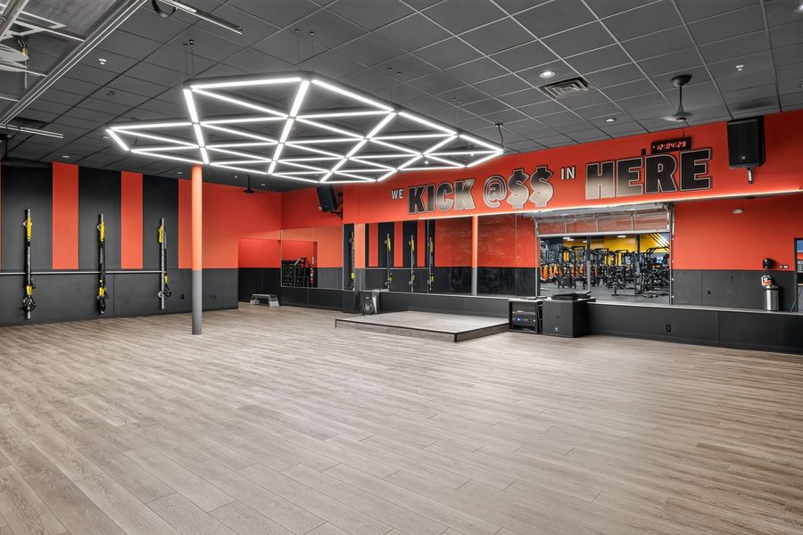 Modern gym interior with orange walls, geometric ceiling lights, hardwood floors, and fitness equipment in the background.