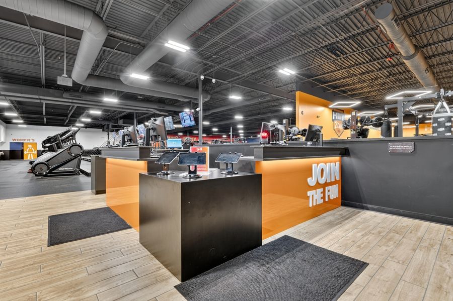 Modern gym reception desk with orange accents and JOIN THE FUN signage, featuring equipment and open workout floor.