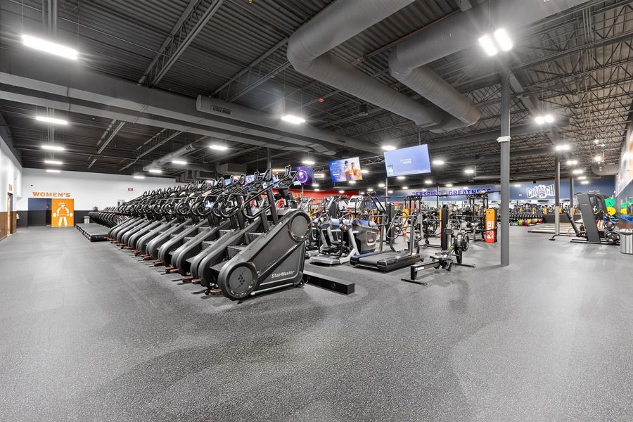 Large modern gym with rows of cardio machines, treadmills, and weight equipment under high ceilings with TV screens.