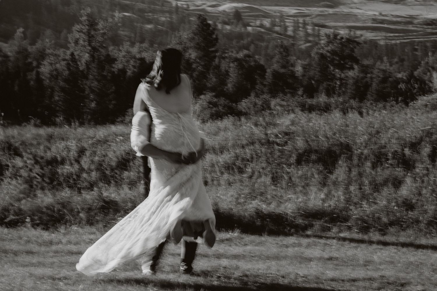 Groom carries bride in white dress through lake water, splashing, with tattoos visible and bouquet in hand.