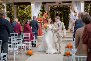 Wedding ceremony aisle lined with white chairs and orange pumpkins at an outdoor venue.