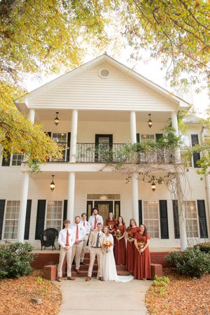 Large white colonial-style house with wedding party in autumn colors standing outside.