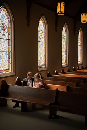 Dark interior of a church sanctuary with stained glass windows casting colored light on wooden pews.