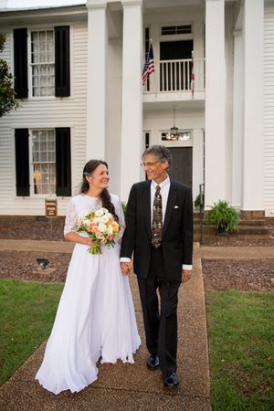 Wedding couple walking together in front of a white columned mansion.