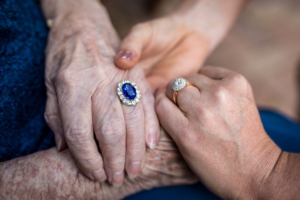 An elderly person and a younger person are holding hands. The elderly person is wearing a ring with a large blue stone surrounded by smaller