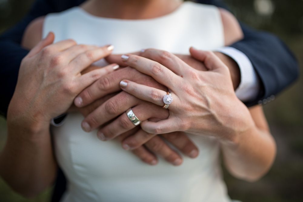 he image shows a close-up of a couple's hands. The woman is wearing a white dress, and both individuals are wearing rings. The man's hands.