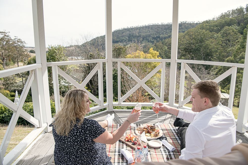 Two people are sitting on a blanket on a porch, enjoying a picnic with food and drinks. They are toasting with glasses The porch has a white