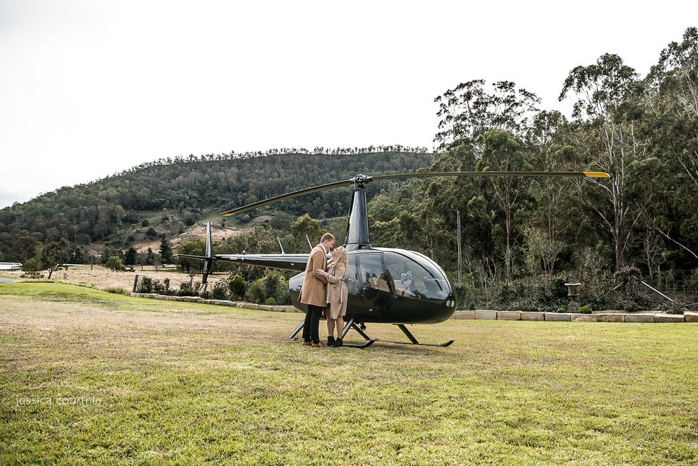A couple is standing in front of a black helicopter on a grassy field They are embracing each other. In the background there are trees hills