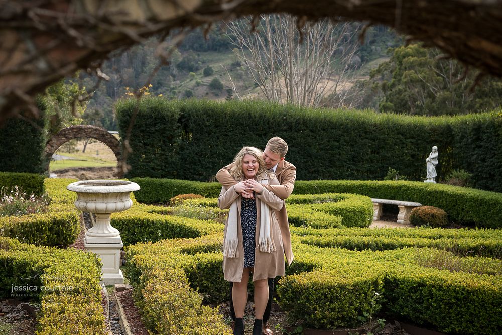 A person is standing behind another person in a garden with neatly trimmed hedges The person in the back is wrapping their arms around front