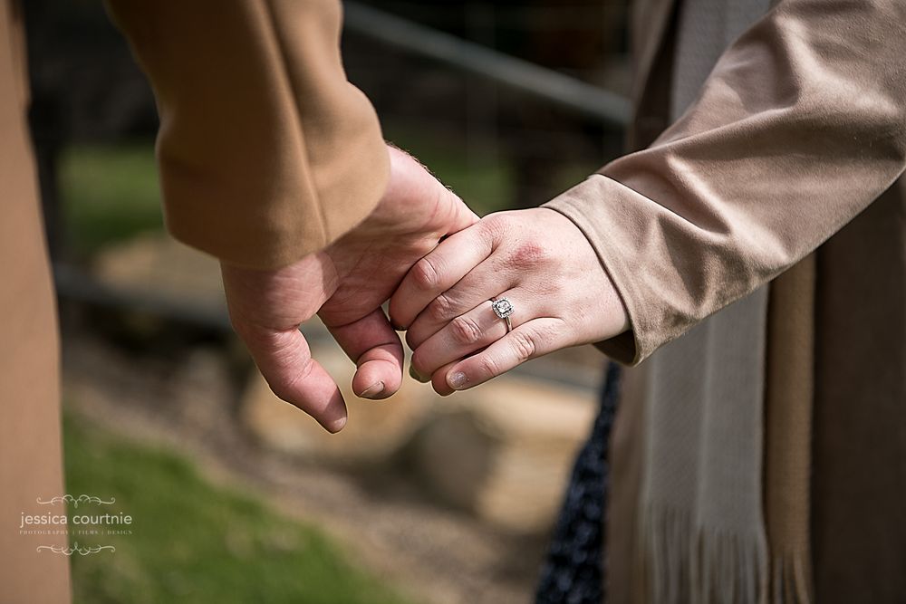 The image shows two people holding hands. One person is wearing a ring on their ring finger. The background is blurred, focusing on the hand