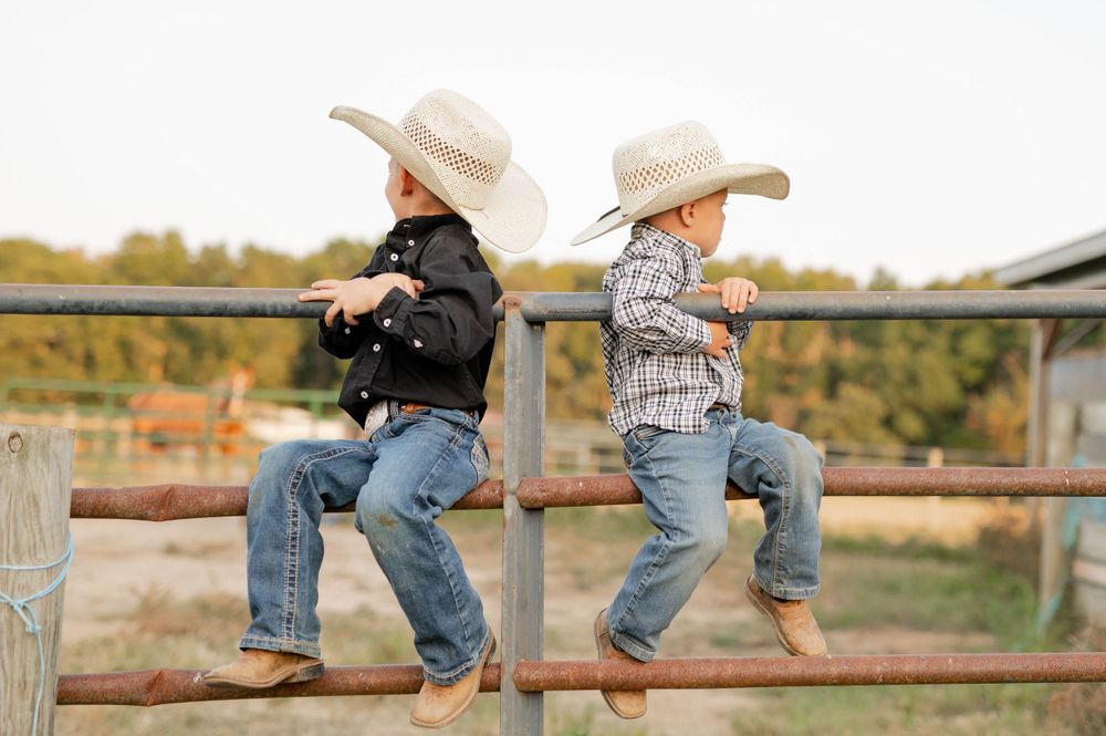 Two children in cowboy hats sit on wooden fence wearing western attire.