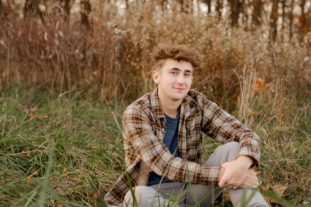 Young person in plaid shirt sits thoughtfully in tall autumn grass at sunset.