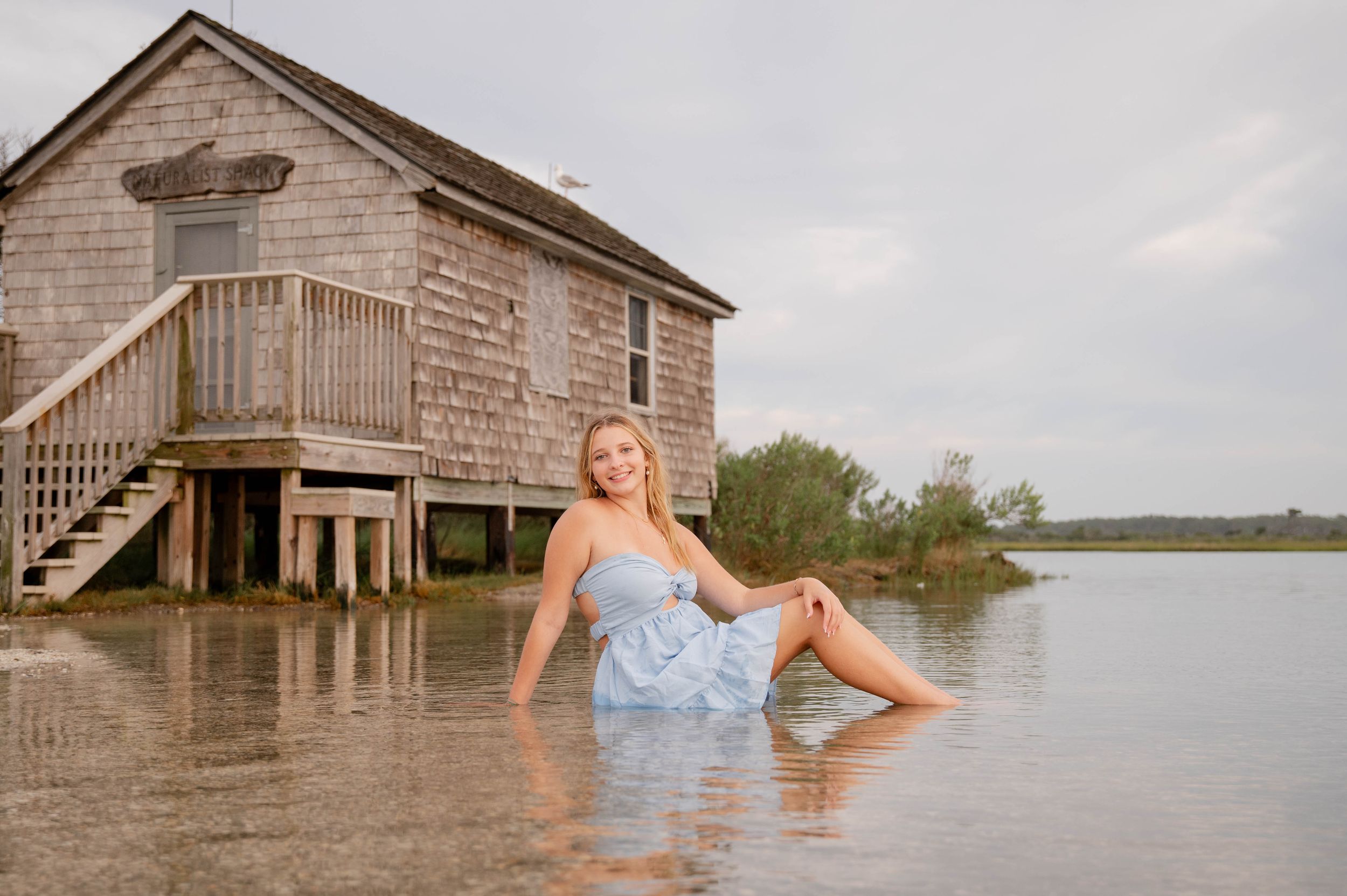 A person in a blue dress poses in floodwaters near a wooden building during a photoshoot.