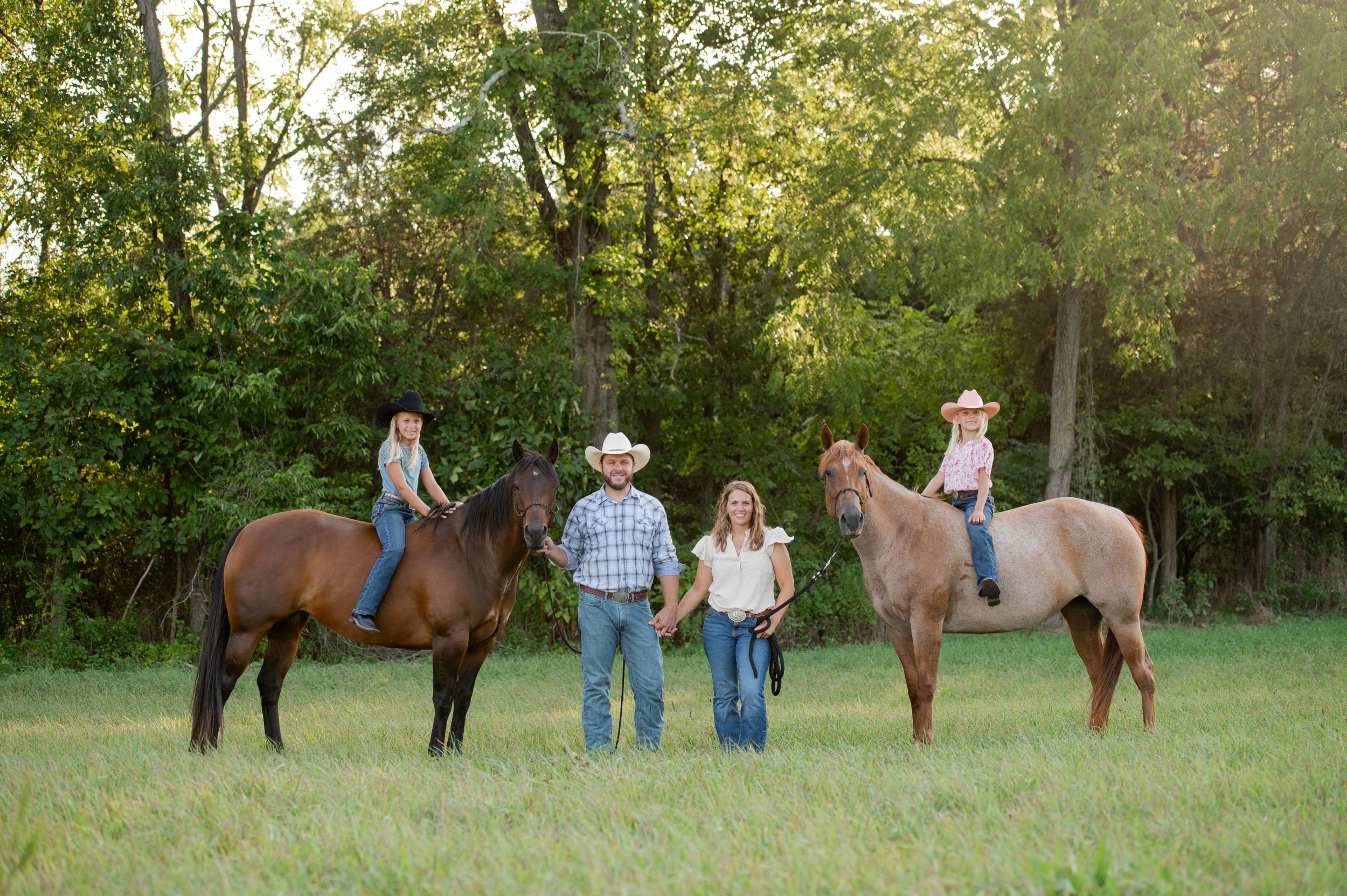 Family group poses with horses in green pasture during golden hour.