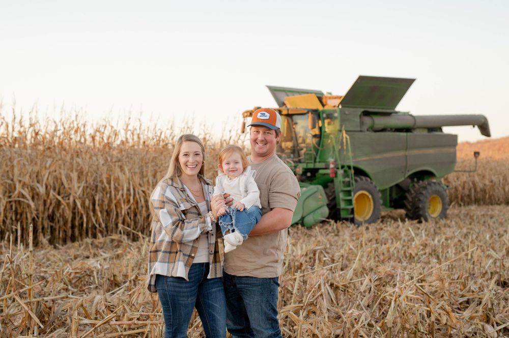 Couple stands in front of John Deere combine harvester in autumn corn field at harvest time.