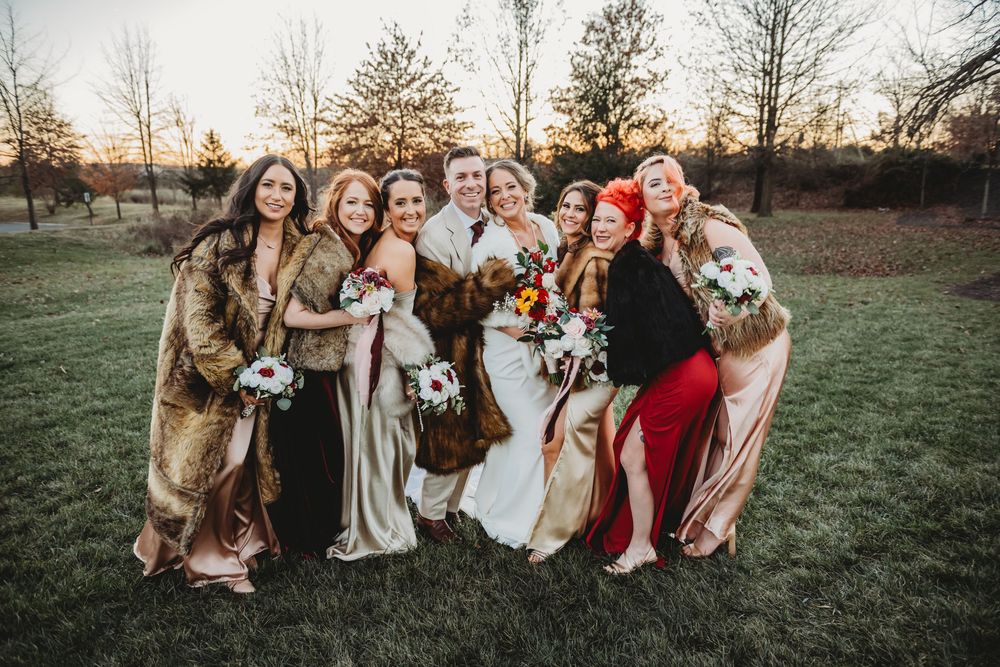 A wedding party group wearing elegant faux fur coats poses together on a grassy field at sunset.