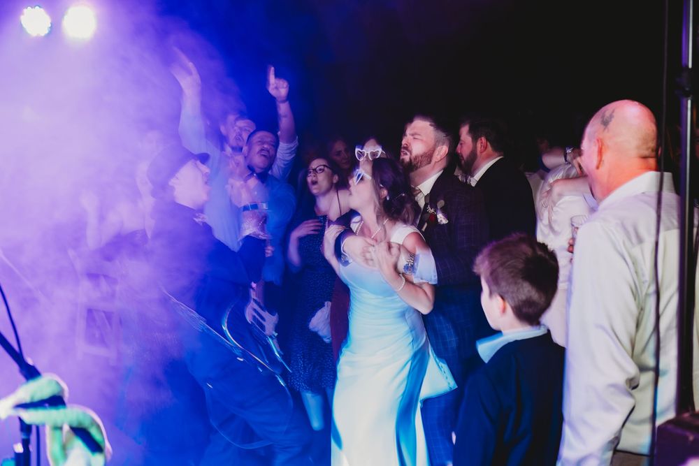 Wedding guests dance and celebrate at a reception with purple lighting effects filling the dance floor.