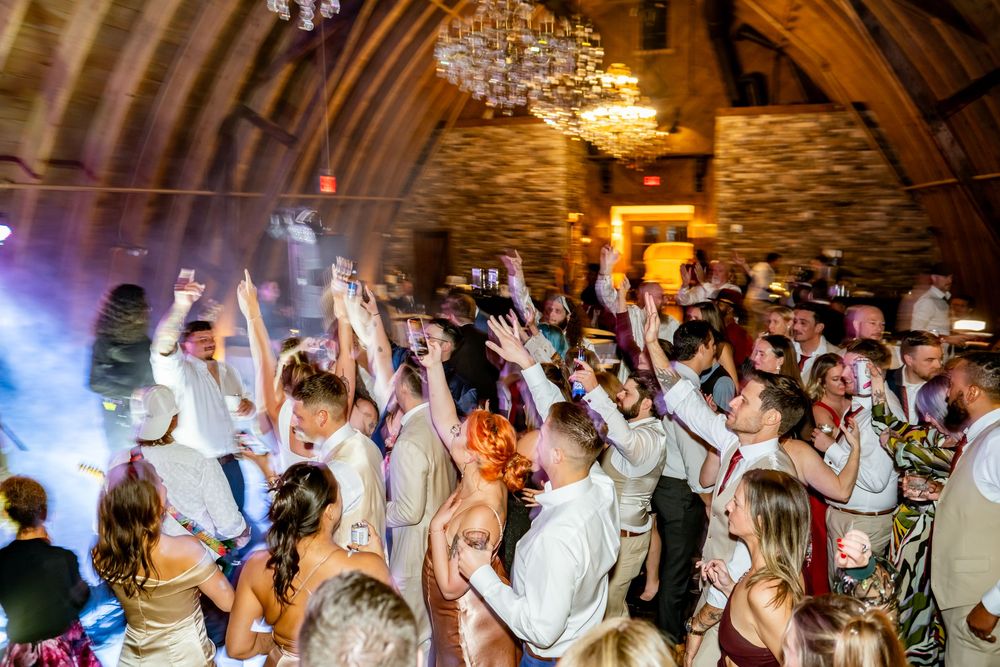 Wedding reception guests dancing and celebrating on a crowded dance floor under chandelier lighting.
