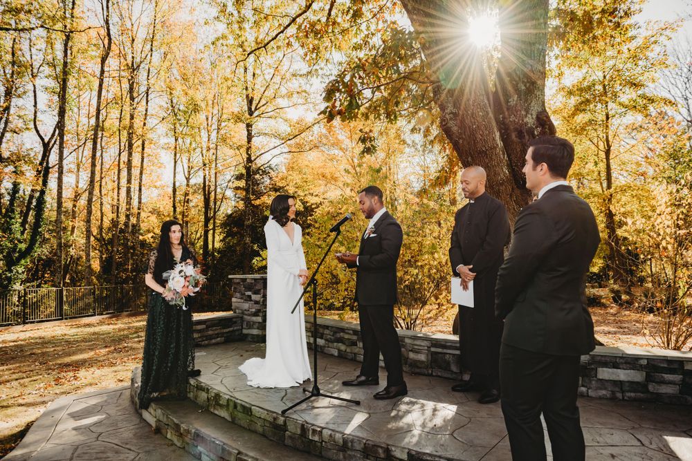 A wedding ceremony takes place under golden autumn trees with sunlight streaming through the branches.