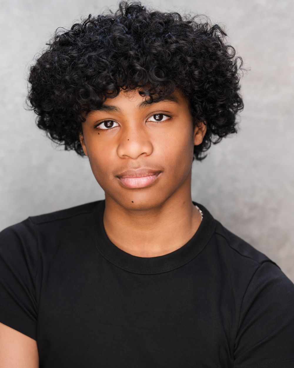 Professional headshot portrait with curly black hair and black shirt against a neutral gray background.
