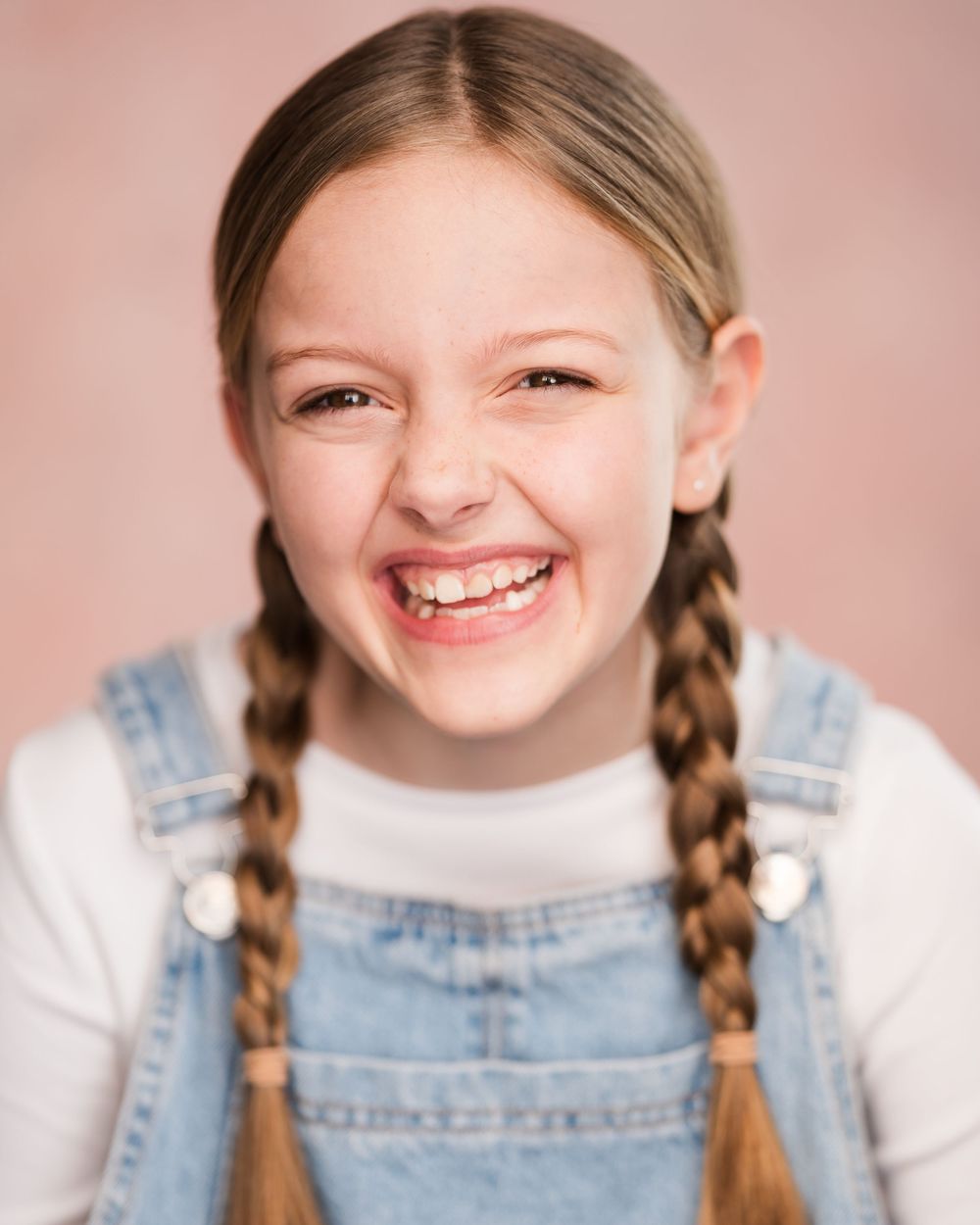 A young person with braided pigtails wearing denim overalls and a white sweater smiles brightly against a pink backdrop.