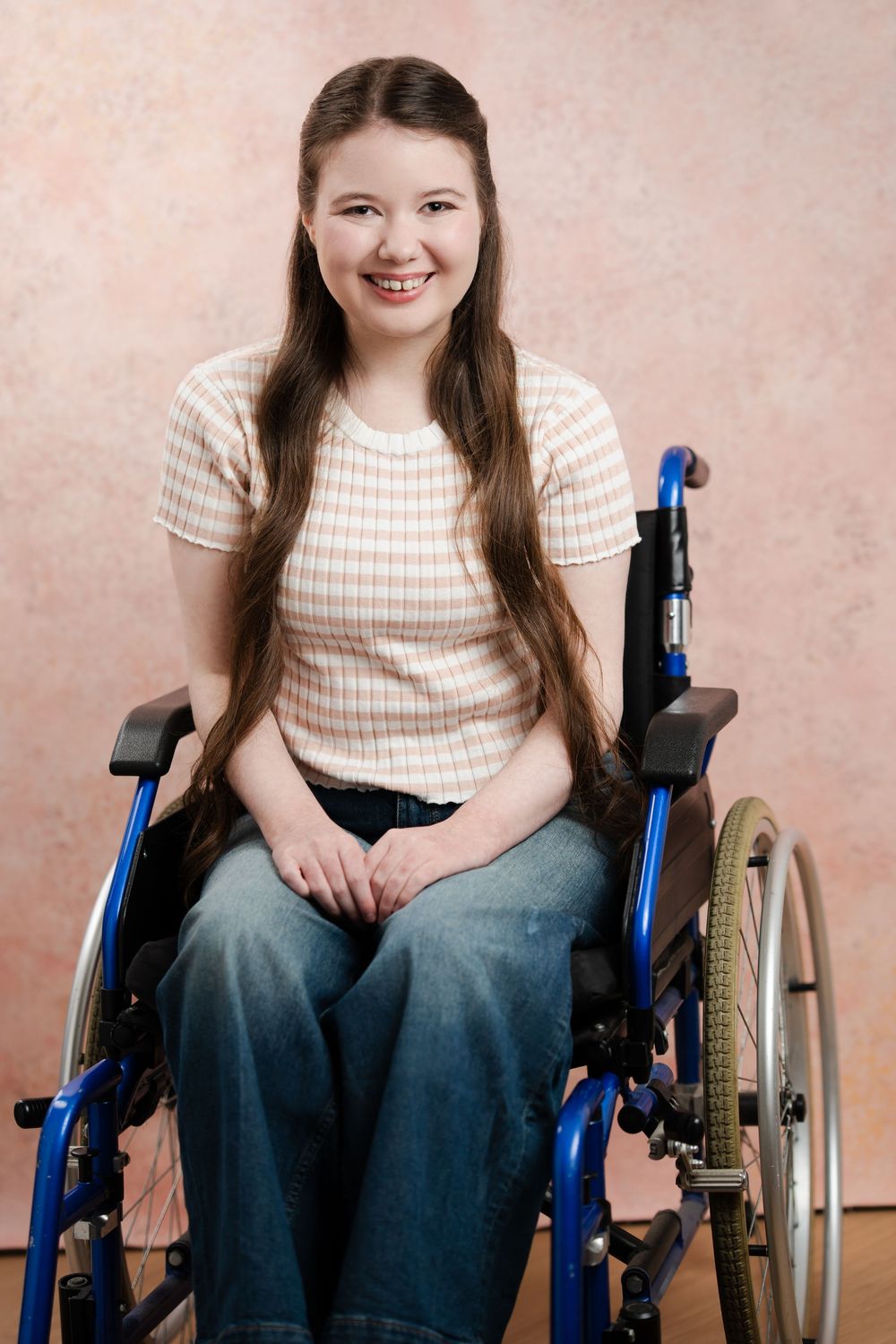 A person in a striped shirt sits in a blue wheelchair against a pink background, smiling warmly at the camera.