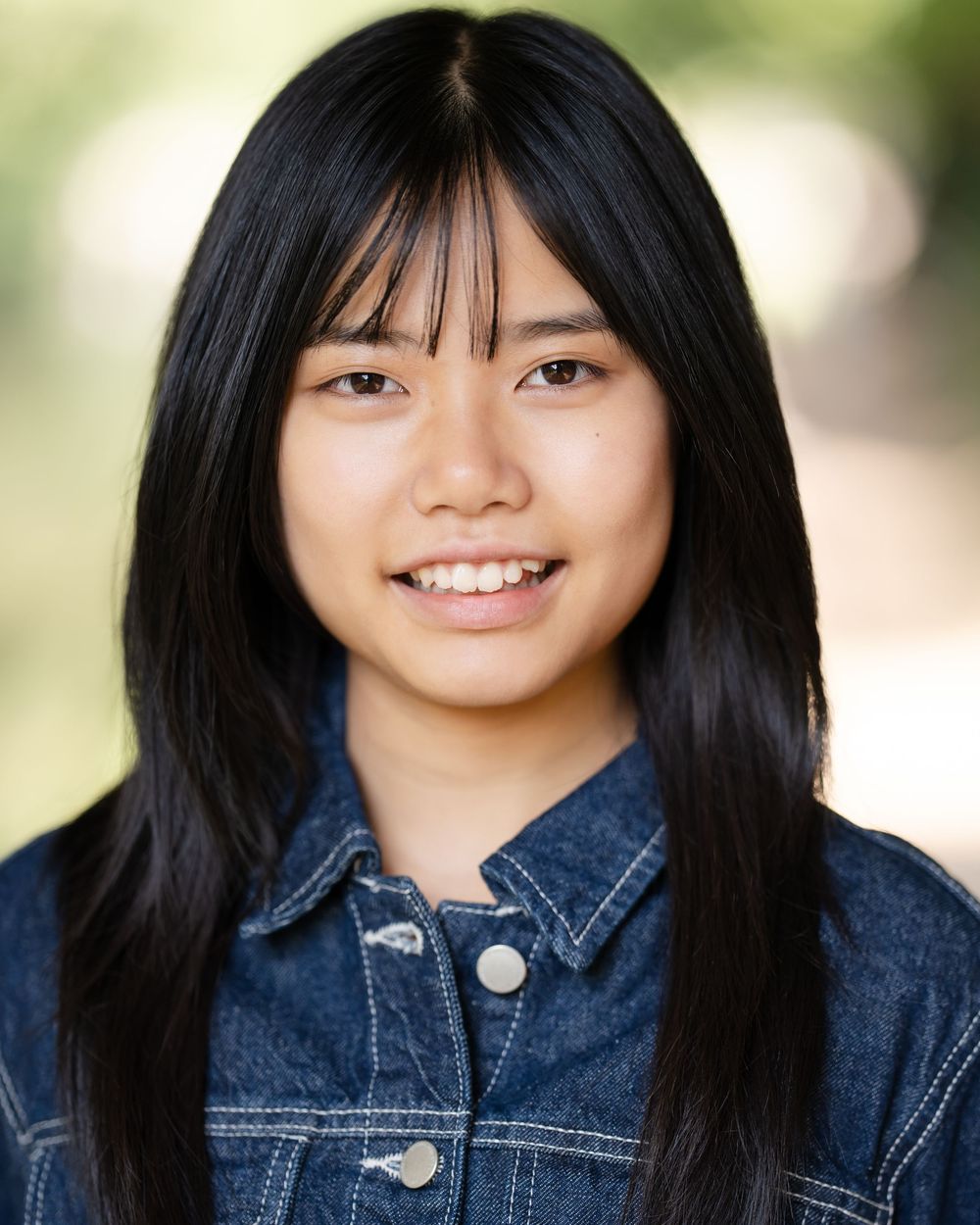 A portrait of someone with long black hair wearing a denim jacket and smiling at the camera against a blurred background.