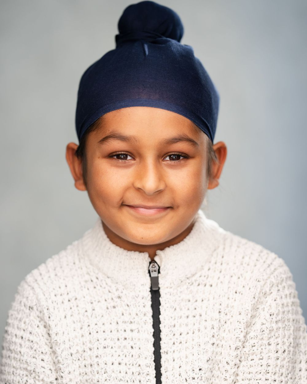 Studio portrait of a child wearing a navy blue turban and white knit sweater against a light background.