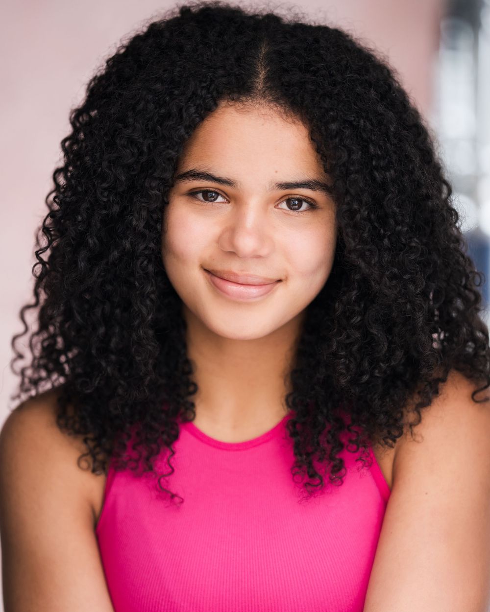 A person with voluminous natural curly black hair wearing a bright pink sleeveless top poses against a light background.
