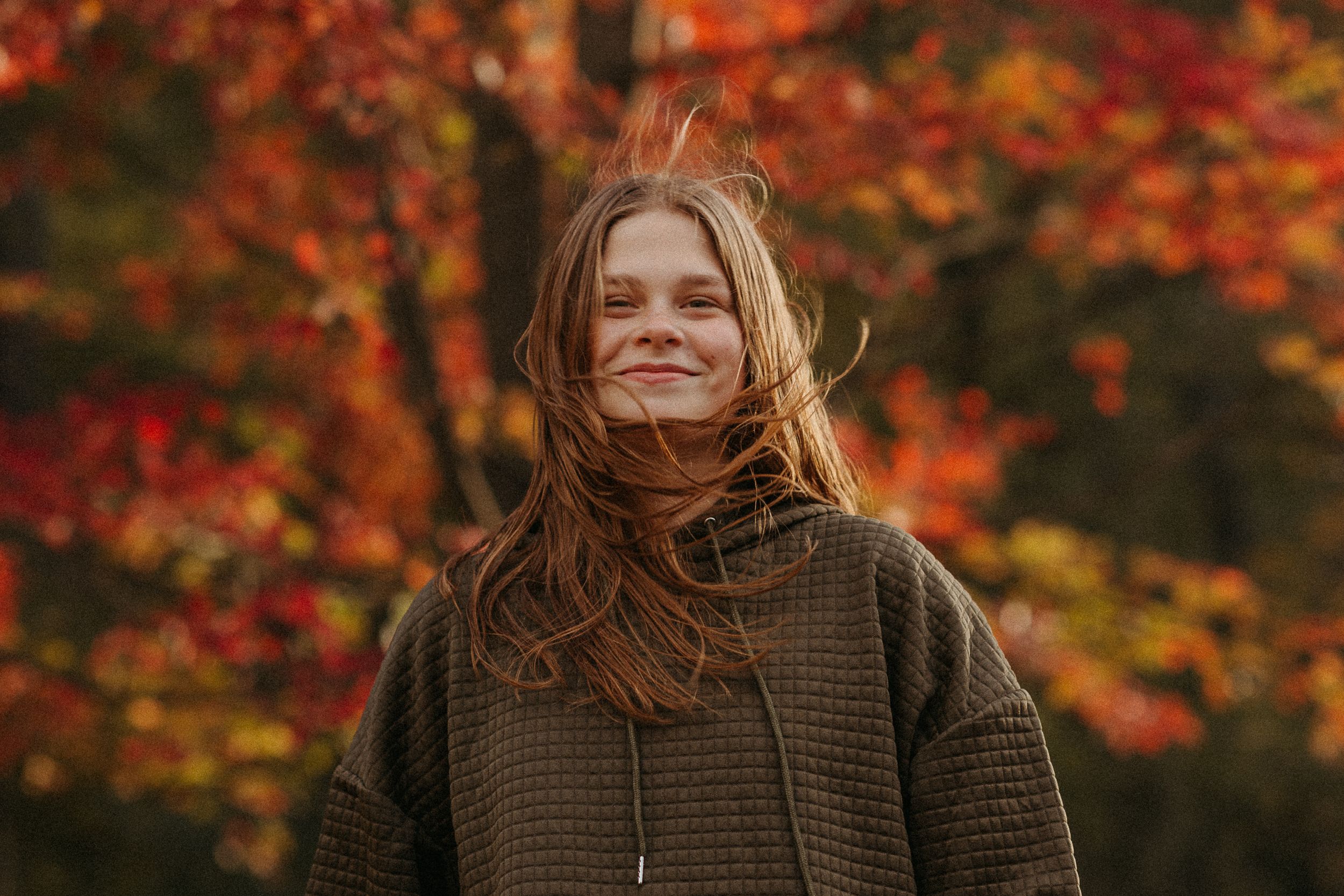 Person in brown sweater smiling against backdrop of vibrant fall foliage.