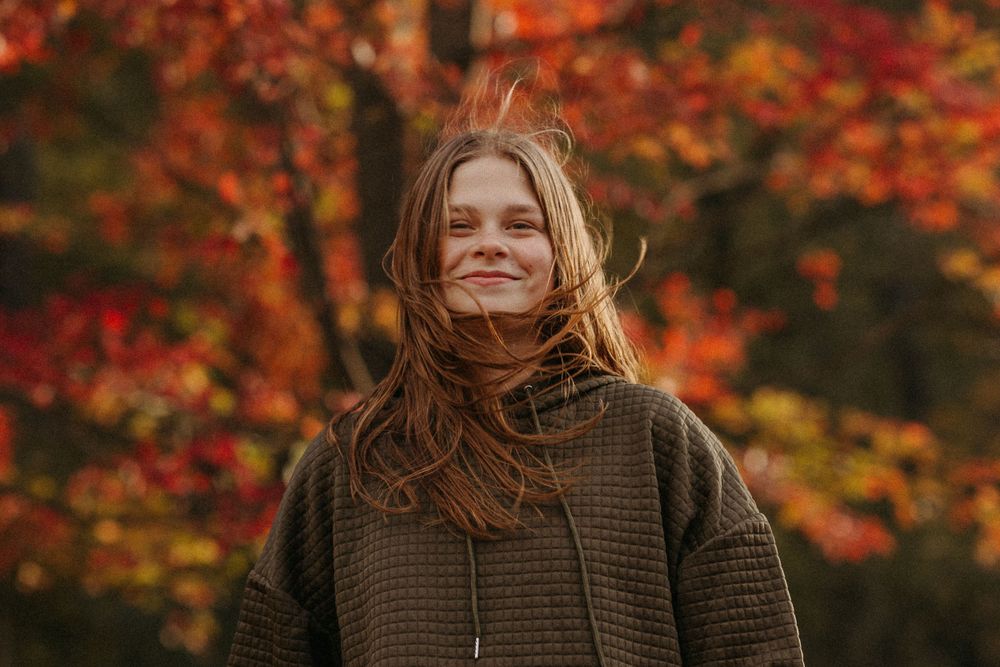 Person in brown sweater smiling against backdrop of vibrant fall foliage.