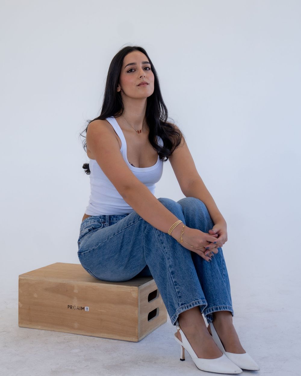 Woman in casual white tank top and jeans sits on wooden box against white backdrop for lifestyle photoshoot.