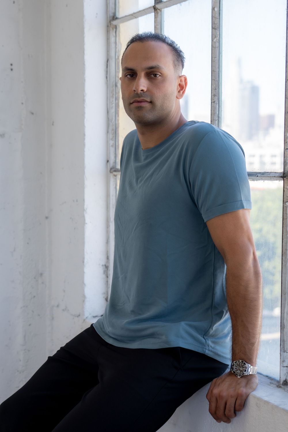 Person in blue t-shirt sits casually by industrial windows with city view.