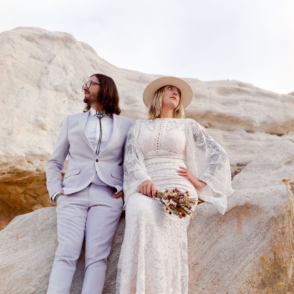 Bride and groom in white lace dress and gray suit pose on rocky desert landscape for boho wedding photo.