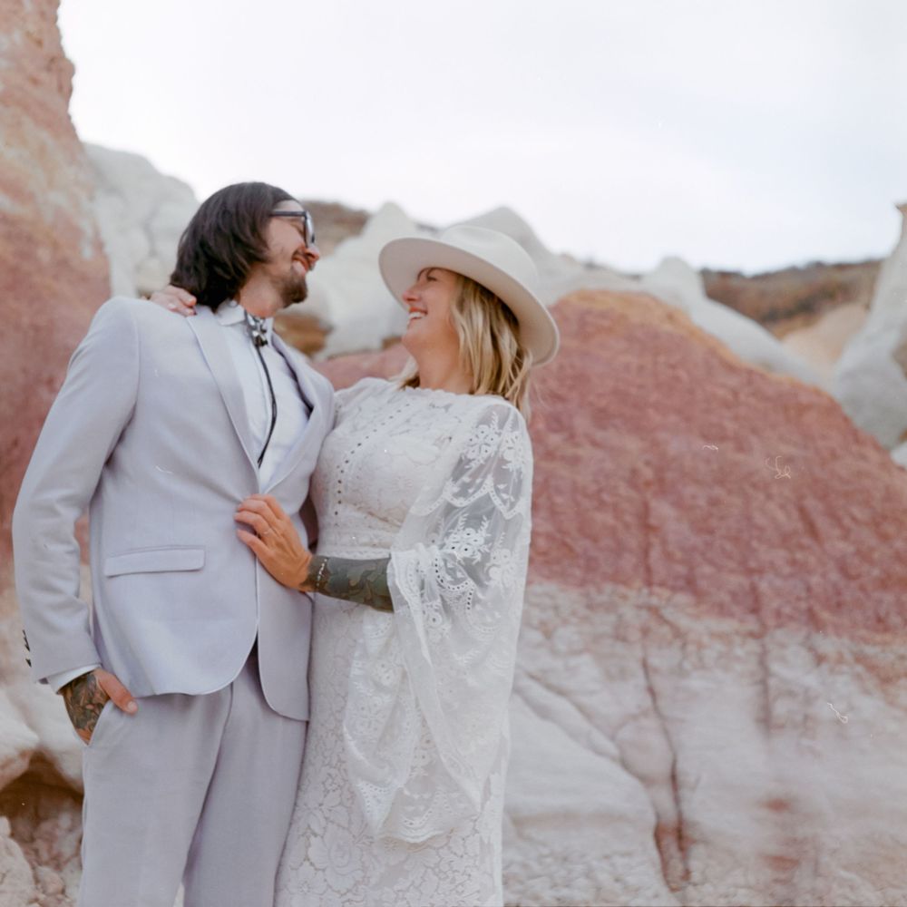 Couple in wedding attire embracing outdoors near colorful red and white rock formations, bride in lace dress and hat.