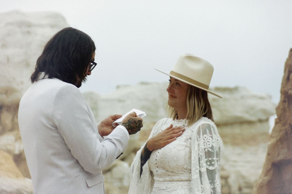 Couple exchanging vows outdoors in a desert setting, bride in white lace dress and hat, groom in light suit.