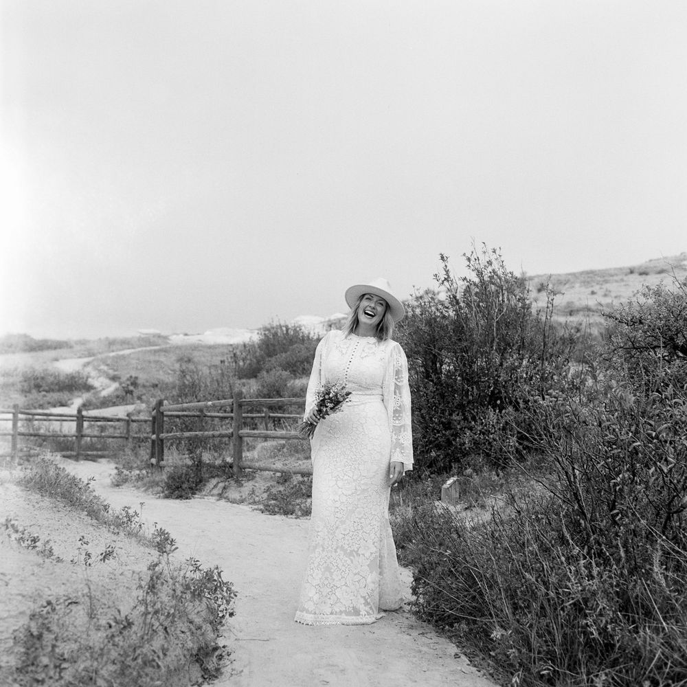 Black and white photo of a bride in lace gown and sun hat holding bouquet on a sandy coastal path.
