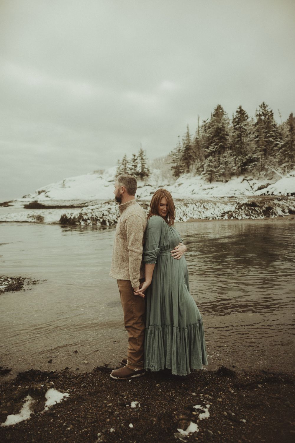 Two people stand together on a rocky beach with snow-covered cliffs and evergreens in the background.